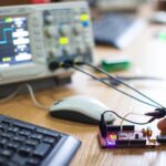 Microcontroller board connected to an oscilloscope on an engineer's desk